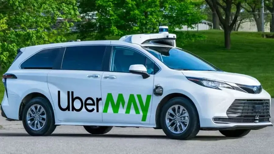 A white Toyota Sienna in Uber and May livery, with a lidar on the roof, parked beside a park