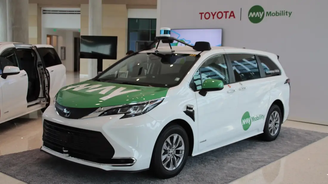 A white and green May Mobility Toyota Sienna on a Toyota and May Mobility display stand, with a lidar on a roof rack